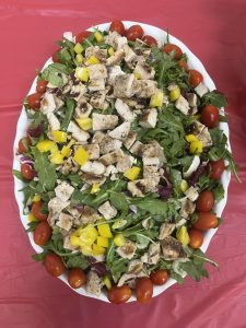 Salad with grilled chicken, yellow peppers, and cherry tomatoes on leafy greens on a red tablecloth.